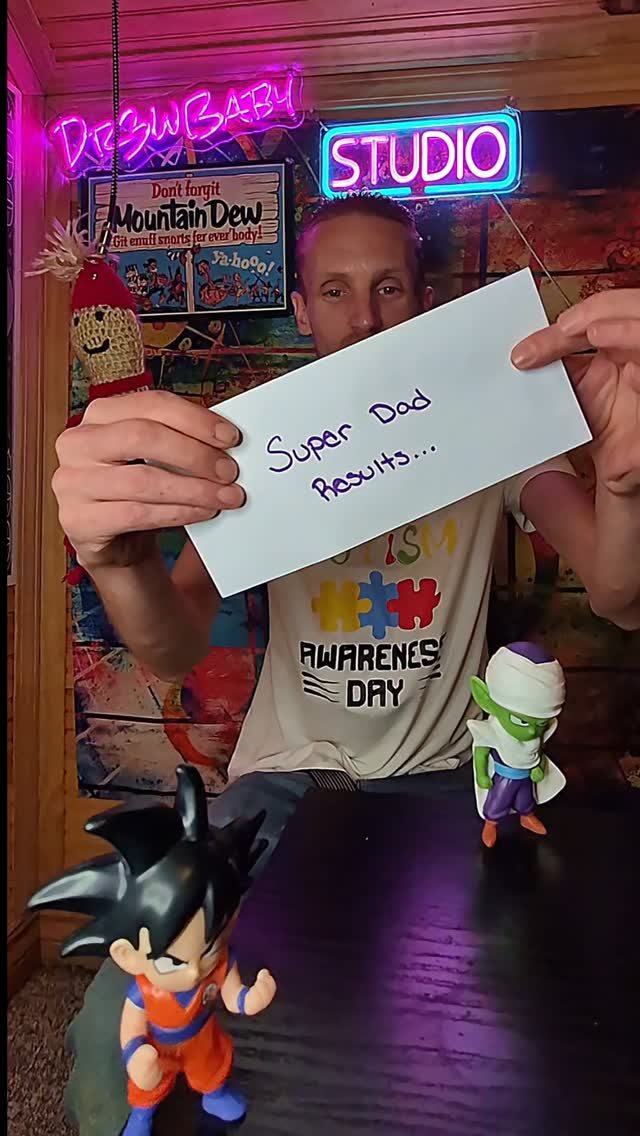Andrew in his home studio holding a Super Dad results card, with Dr3wBaby and Studio neon signs behind him